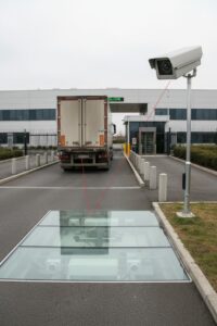 Ein LKW fährt über ein im Boden eingelassenes Kamerasystem zur Fahrzeugunterbodenkontrolle, während eine Überwachungskamera auf einem Mast den Vorgang erfasst.